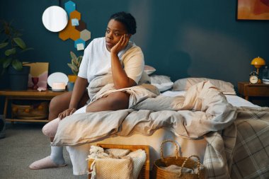 Düşünceli bir ifadeyle, modern dekor elementleri ve düzgün bir yatağı olan rahat bir yatak odasında yatağın kenarında oturan bir kadın.