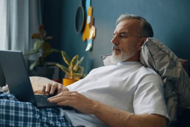 Beyaz sakallı yaşlı bir adam dizüstü bilgisayar kullanarak yatakta yatıyor. Çevrelerinde bitkiler, duvar süslemeleri ve rahat bir yastık yer alıyor.