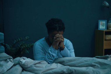 Elleri bağlı bir şekilde, arka planda bitkiler ve yanında ahşap mobilyalarla loş bir odada dua eden bir kadın.