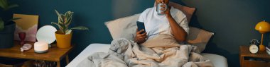 Elinde akıllı bir telefonla yatakta kahve içen kişi. Etrafı bitkiler, lamba ve diğer dekorasyon malzemeleriyle çevrili. Huzurlu bir sabah atmosferi yaratıyor.