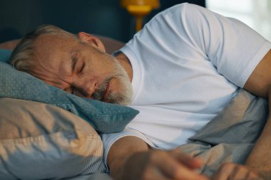 Yaşlı bir adam yatakta uzanıyor, başını yastığa koyup rahatça uyuyor, doğal ve huzurlu bir ifade sergiliyor, huzur verici bir huzur ortamı yaratıyor.