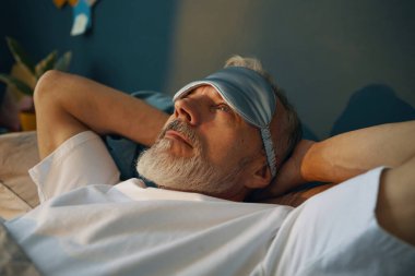 Yatakta uzanan yaşlı adam, uyku maskesi takıyor ve sakin görünüyor. Arka planda bitkiler ve rahat atmosferli minimalist dekorlar görünüyor