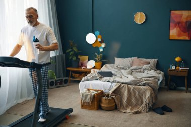 Sıcak ve renkli yatak odasında koşu bandında yürüyen yaşlı bir adam susuzluk için su şişesi tutuyor. Yatak odasında modern dekor ve doğal ışık ile düzenli bir alan var.