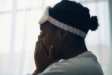 Beyaz bir uyku maskesi takan Afro-Amerikalı kadın elleriyle yüzünü örtüyor, sakin ve içe dönük görünüyor.