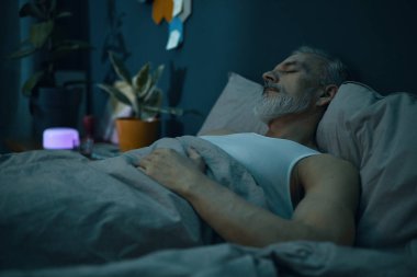 Gri saçlı yaşlı adam rahat bir yatak odasında uyuyor. Bitkiler ve yumuşak ışıklı yatak, rahatlatıcı bir atmosfer yaratıyor.