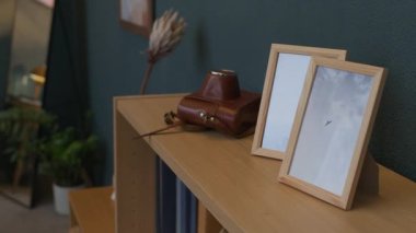 Klasik deri kamera çantası, iki çerçeveli fotoğraf ve minimalist oturma odasındaki ahşap rafta kitaplar.
