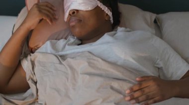 Genç siyahi kadının yatakta uyanması, uyku maskesini çıkarması, sabah evde gülümsemesi ve esnemesi.
