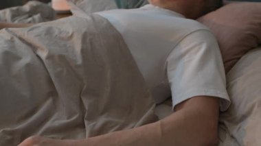 Yatakta uzanmış, evde uyuyan rahat yaşlı bir adamın, yakınlardaki komodinin üzerinde kapalı bitkiler ve lambayla çekilmiş bir fotoğrafını çek.