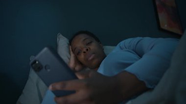 Genç siyahi kadın, gece uykusuzluk çekerken, elinde akıllı telefon ve sosyal medyada kaydırma yaparak karanlık odada uzanıyor.