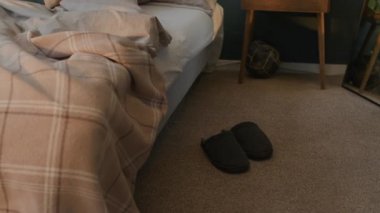 Yastıklı ve battaniyeli yapılmamış yatağı, halı döşemesinin yanındaki koyu renkli terlikleri, rahat yatak odasındaki ahşap komodinin yanına yatır.