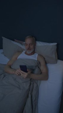Beyaz bir adamın yatakta battaniyenin altında yattığı dikey görüntüsü, gece geç saatlerde loş bir odada akıllı telefondan gülümseyerek kayıyor.
