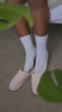Pijama ve çorap giymiş, ayaklarını terlik giymiş, sabahları yataktan çıkarken esneyen ve gülümseyen genç ve pozitif siyahi bir kadının dikey poziyonu.