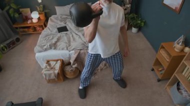 Pijama ve terlik giymiş üst düzey bir adamın yatak odasında durup, kettlebell salıncakları sallarken ve evde egzersiz yaparken tek koluyla yaptığı yüksek açı görüntüsü.