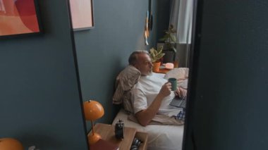 Ayna yansıması, kıdemli bir adamın rahat bir odada yatakta dinlenmesi, kahve içmesi ve sabah evde dizüstü bilgisayarla internette sörf yapması.