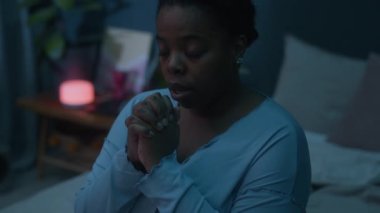 Pijamalı siyah bir kadın akşamleyin loş bir odada elleri bağlı ve gözleri kapalı dua ediyor.