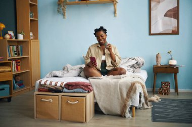 Yatakta oturmuş gülümseyen bir kadın, elinde cep telefonu, sıcacık bir dekor ve sıcak aydınlatmayla çevrili. Rahat ve rahat bir atmosfer gösteriliyor