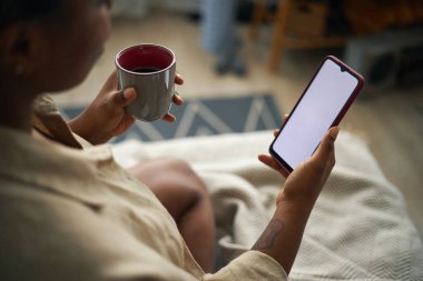 Akıllı telefona bakarken elinde kahve fincanı tutan kadın oturma odasında dinleniyor. Ekrana odaklanmış, kesintisiz boş anın tadını çıkarıyor.