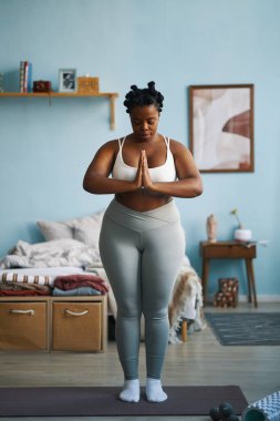 Aktif giysi giyen Afrikalı Amerikalı kadın ellerini dua pozisyonuna koyarak farkındalık yogası yapıyor, yatak odasındaki rahatlamaya odaklanıyor.