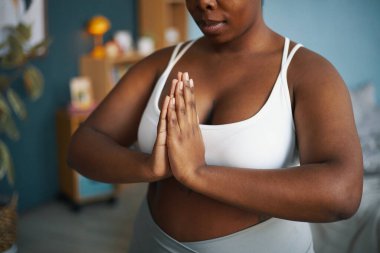 Afro-Amerikalı bir kadın ellerini dua pozisyonunda uzatarak meditasyon yapıyor, içeride duruyor, sakin ve rahat hissediyor, iyi aydınlatılmış bir odada.