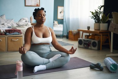 Afrika kökenli Amerikalı bir kadın yoga minderinin üzerinde bağdaş kurarak büyük pencereden süzülen yumuşak doğal ışıkla dinlenme odasında meditasyon yapıyor.