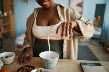 Afro-Amerikalı kadın, mutfak ortamında mobilya ve dekorla çevrili seramik bardağa badem sütü dökerken gülümsüyor.