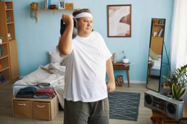 Beyaz saç bandı takan ve beyaz gömlek giyen bir adam yatak odasında müzik dinliyor. Etrafı dağınık eşyalar ve dekorlarla dolu.