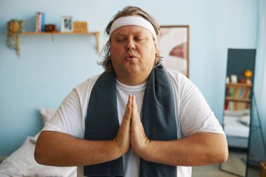 Elleri dua pozisyonunda orta yaşlı bir adam huzur içinde meditasyon yapıyor. Çerçevelenmiş resim, raflar, yatak ve kitaplar rahat bir ortam yaratıyor.