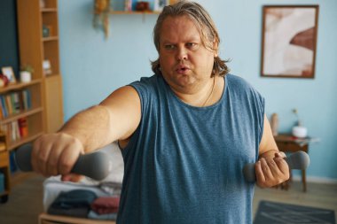 Kafkasyalı bir adam içeride dambıllarla egzersiz yapıyor odaklanmış görünüyor. Kitaplar ve dekorasyonlarla dolu oturma odası ayarları rahat bir atmosfer ekliyor.