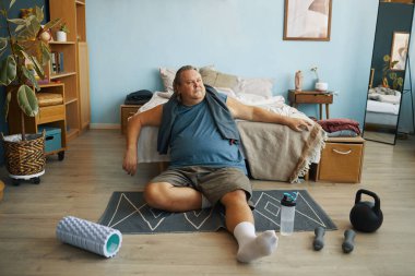 Konforlu bir odada egzersiz yaptıktan sonra yoga minderinde dinlenen ve spor sonrası rahatlamayı yansıtan bir adam.