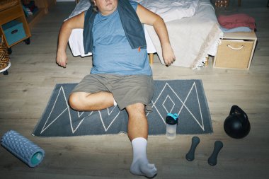 Yoğun bir egzersizden sonra dinlenen kişi, ahşap zeminde egzersiz ekipmanlarıyla çevrili.
