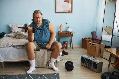 Ev ortamında yatağın yanındaki bankta oturan orta yaşlı bir adam dambıl kaldırıyor.