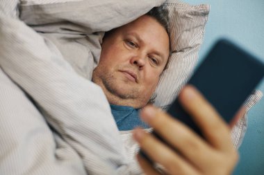 Rahat bir adam yatakta uzanmış, akıllı bir telefona bakıyor.