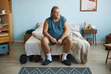 Adam rahat oturma odasında kanepede oturuyor, elleri sımsıkı bağlı ve dalgın görünüyor. Ev içinde raflar, kitaplar ve yerdeki ağırlıklar vardır.