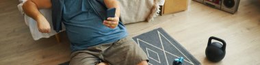 Akıllı telefon, kettlebell ve su şişesiyle yakınlarda rahat bir yaşam alanında yerde uzanan biri. Bir el akıllı telefonu tutuyor.