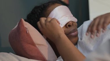 Genç siyahi kadının yatakta uyandığı, uyku maskesini çıkardığı ve sabah erkenden yastığa koyduğu yakın plan.