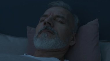 Endişeli yaşlı adamın yataktaki kabustan uyanıp yüzünü ovalarken ve gece boyunca sakinleşmeye çalışırken yakın plan çekimleri.