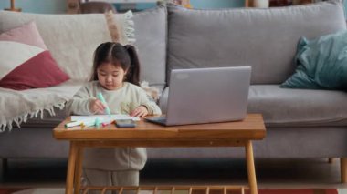 Orta ölçekli mutlu Asyalı bebek masadaki bilgisayarı ve rahat evdeki akıllı telefonu kağıt üzerine çiziyor.