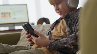 Gülümseyen beyaz çocuk akıllı telefon kullanırken rahat koltukta dinleniyor, evde dijital eğlencenin tadını çıkarıyor, arka planda bilgisayar ekranı var.