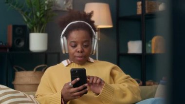 Sarı kazaklı, gülümseyen siyah kadın müzik dinliyor ya da kablosuz, kulaklıklı canlı yayında rahat oturma odasındaki kanepede akıllı telefonunu kullanıyor.