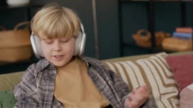 Teknolojiden anlayan beyaz çocuk, akıllı telefon kullanarak evdeki yumuşak kanepede dinleniyor, kablosuz kulaklık takıyor, akıllı kulaklıklar takıyor, modern dijital hayata dalmış.