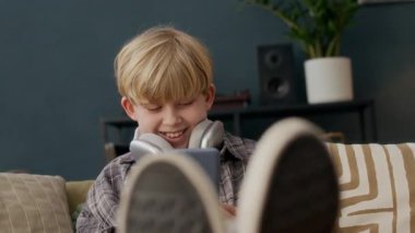 Ekose gömlekli neşeli beyaz çocuk ayaklarını kaldırmış, akıllı telefon ekranına gülümsüyor, kulaklıkları boynunda, evde dijital eğlencenin keyfini çıkarıyor.
