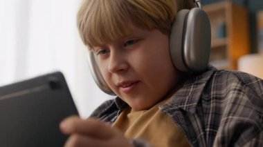 Evdeki tabletinde oyun oynarken duygusal hayal kırıklığını gösteren kablosuz kulaklıklı sinirli beyaz çocuğun yakın çekimi.