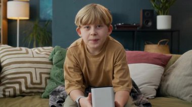Teknoloji meraklısı beyaz çocuk elinde akıllı hoparlörle kameraya hitap ediyor, sonra rahat, modern oturma odası ortamında koltuğa yaslanıyor.