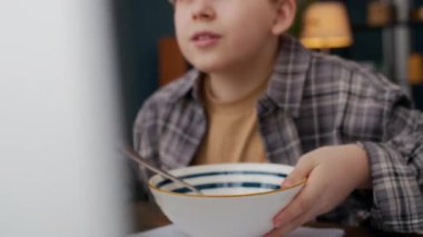 Günlük ekose gömlekli beyaz bir çocuk, bilgisayarında oyuna dalmış, mısır gevreği kasesine süt ekliyor ve yemek yemeye başlıyor, modern apartmandaki çalışma masasında kahvaltı yapıyor.