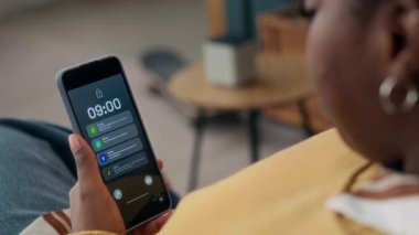Sarı kazaklı genç siyahi kadının akıllı telefonuyla akıllı ev tebligatlarını izlediği, elektrikli süpürge ve akıllı hoparlörler gibi cihazlarla ilgili güncellemeleri sabah 9 'da sergilediği omuz üstünden çekim.