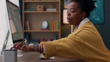 Yan görünüm, sarı kazaklı siyahi kadın masaüstü bilgisayarı üzerinde çalışırken akıllı hoparlörleri kullanmak için uzanıyor.