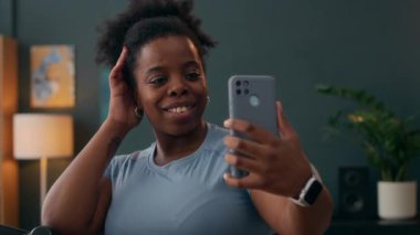 Aktif giyimli ve akıllı yüzlü gülümseyen genç siyahi kadın evdeki koşu bandındaki antrenmanına başlamadan önce akıllı telefonuyla hızlı bir selfie çekiyor. Fitness, eğlence ve teknoloji karışımı.