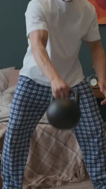 Ev elbiseli yaşlı bir adamın dikey olarak yatak odasında durduğu ve günlük antrenman sırasında kettlebell salıncakları yaptığı sahne.