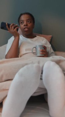 Yatakta rahat bir şekilde oturan genç siyahi kadının dikey görüntüsü, elinde kupa tutarken ve evde hoparlörle konuşurken.