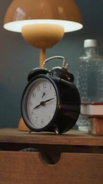 Analog çalar saatin dikey yakın çekimi, kitap yığını, su şişesi ve lamba ahşap komodinin üzerinde.